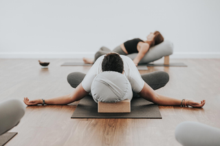 A person in a white shirt and gray pants holds a forward bend yoga pose on a dark yoga mat, using a light gray headrest pillow and a wooden block. Another person is visible in the background on a yoga mat in a bright studio with a wooden floor.