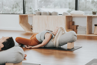 A woman in a yoga pose lies on a black mat using a large gray bolster pillow with a wooden block support in a bright, modern yoga studio.