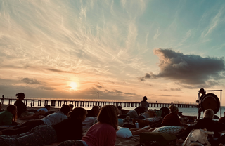 A sunset scene at a beach or pier with many people sitting and standing, silhouetted against a glowing sky with wispy clouds and a large dark cloud on the right.